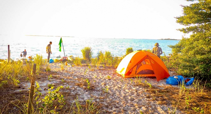 An orange tent rests on the sand of a beach. There are a few people around, and vast blue ocean in the background. 